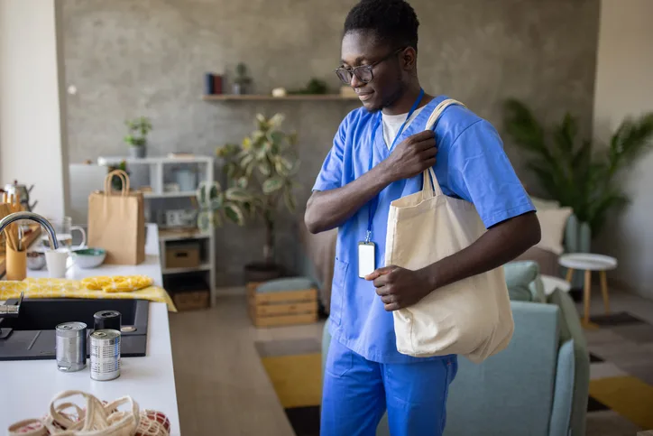A young African American male nurse wearing medical scrubs bringing groceries to his apartments kitchen