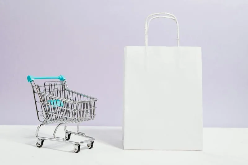 A miniature shopping cart beside a blank white gift bag, representing the importance of thoughtful purchasing decisions when ordering custom swag.