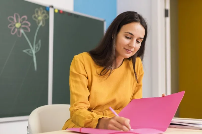 Teacher writing in pink folder at desk with chalkboard behind