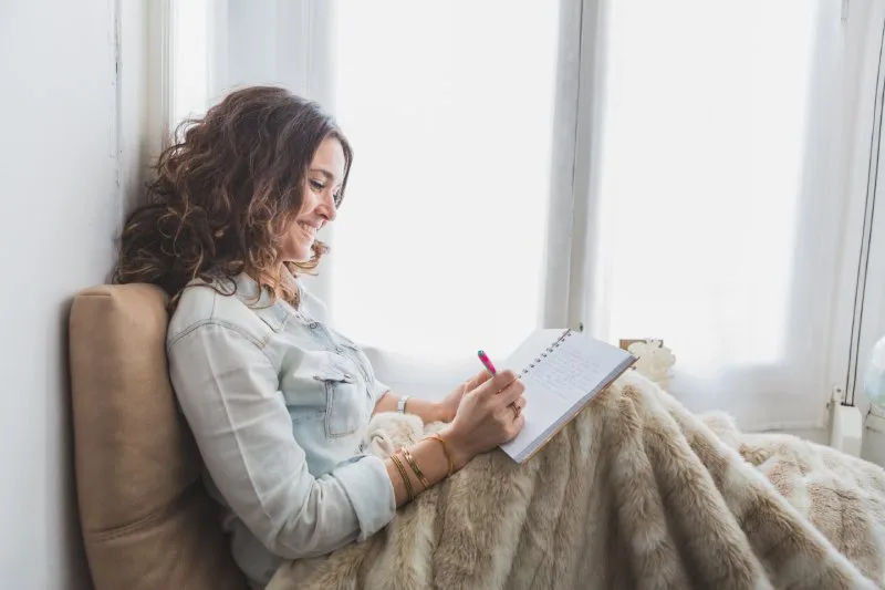 Smiling woman sitting by window with blanket, writing in journal, mindfulness employee wellness gifts idea.