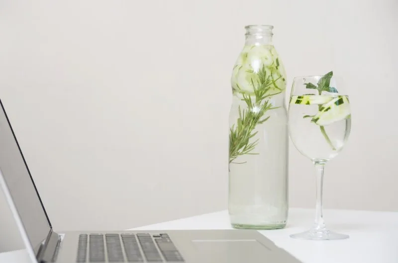 Glass bottle with cucumber and herb-infused water beside laptop, hydration-focused employee wellness gifts idea.
