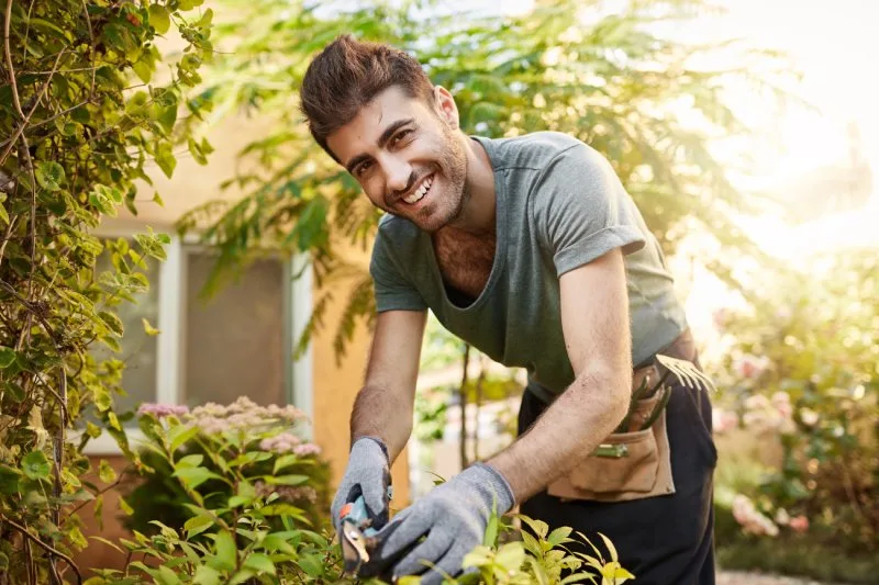 Smiling man wearing gloves trimming garden plants outdoors, personal growth employee wellness gifts promoting relaxation.