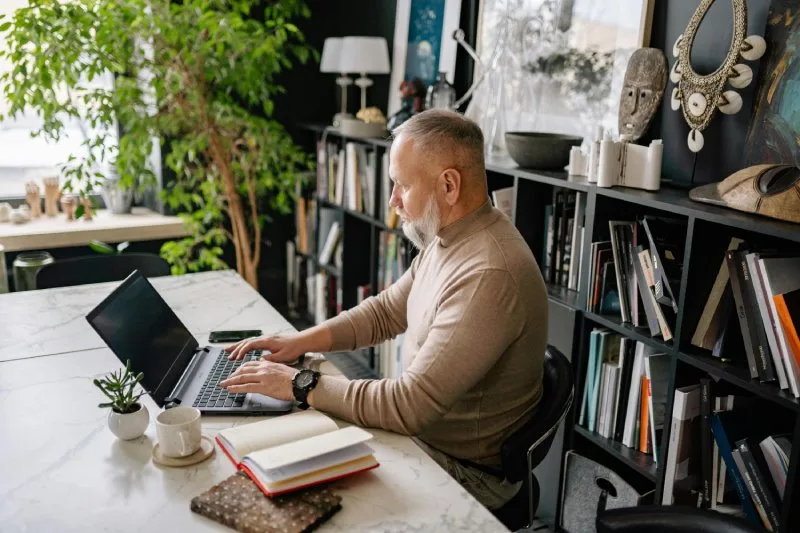 Older man working on laptop at desk with notebooks and coffee, workplace employee wellness gifts.